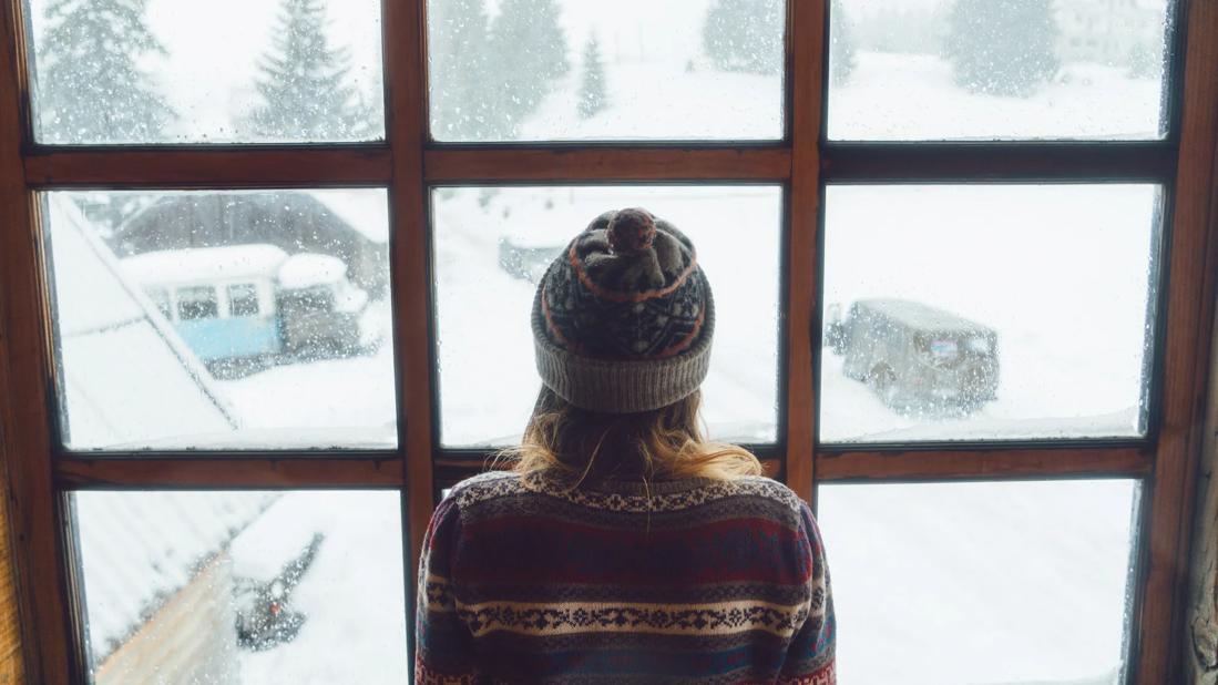 Person in sweater and knit hat facing window, with winter, snowy yard outside