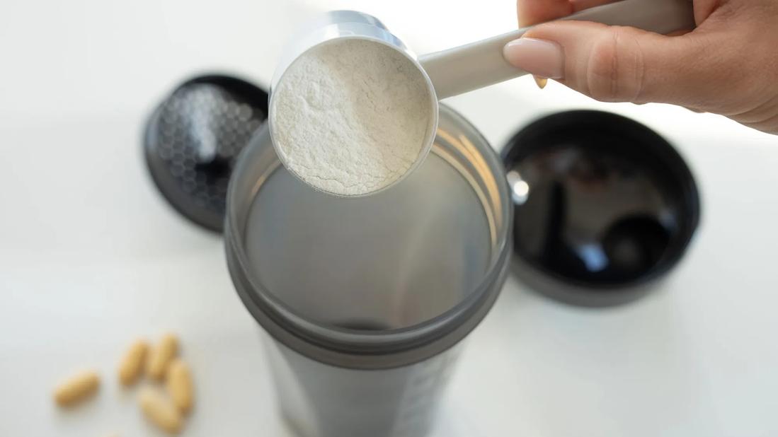 Hand pouring scoop of supplement powder into shaker, with blurry pills on the counter