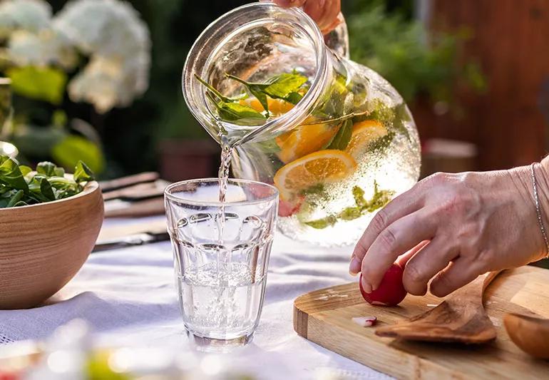 Person pouring a pitcher of cold water with oranges and herbs into a glass cup