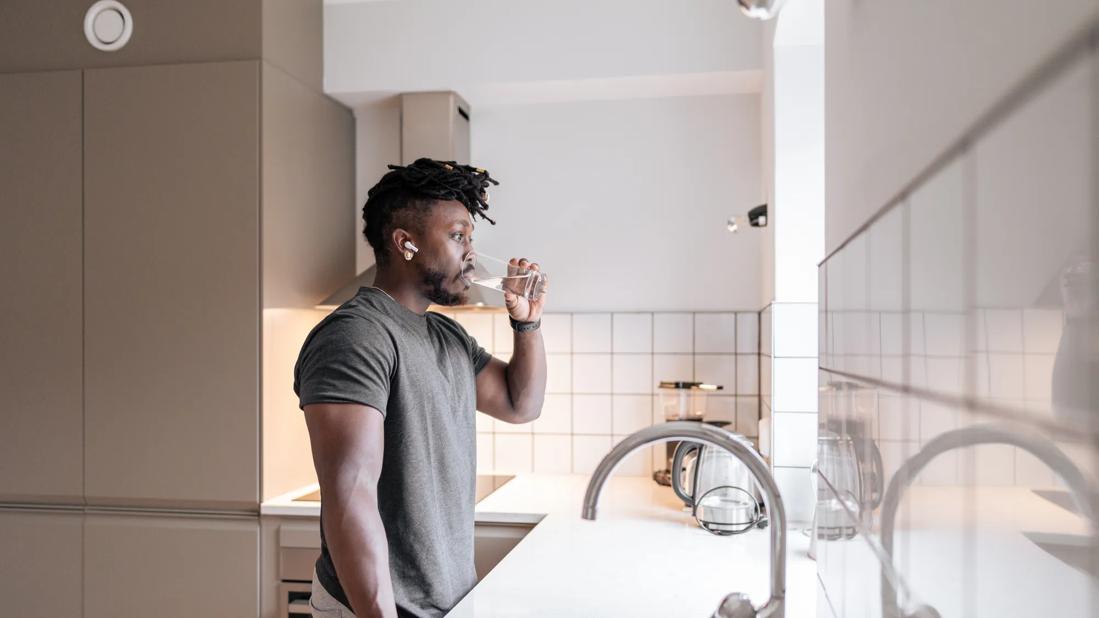 Person with earbud in standing at kitchen sink drinking a glass of water