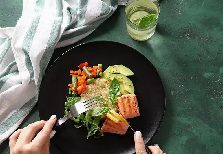 Person cutting salmon and fresh vegetables on dinner plate.