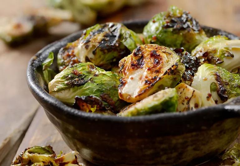 A closeup of roasted brussel sprouts in a brown bowl with more brussel sprouts in background.