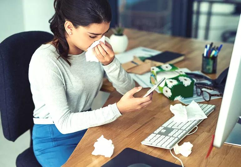 Woman sitting at computer wiping nose and surrounded by used tissues