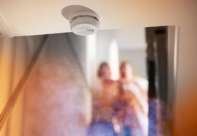 Closeup of a smoke swirling up to a smoke detector, with a couple visible in the background