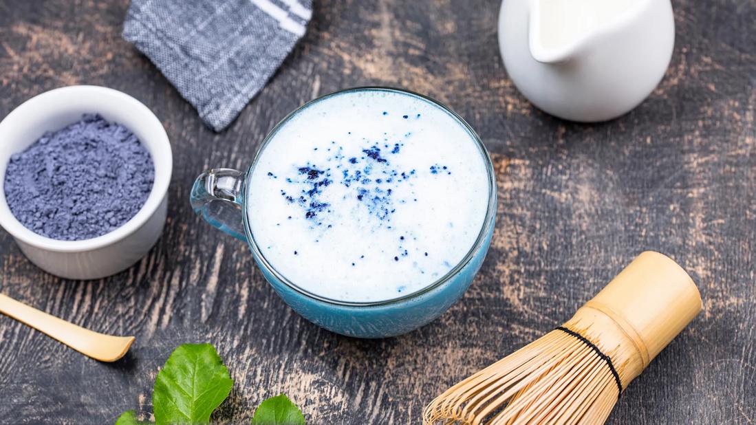 Blue matcha powder and matcha-making equipment surround a glass mug of blue matcha tea