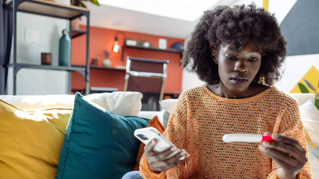 Female sitting on couch looking at a pregnancy test stick, holding cell phone