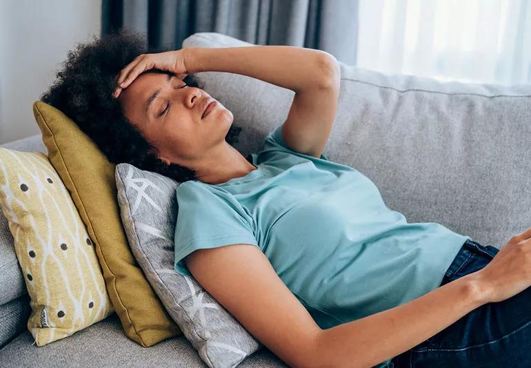 Person lying on couch holding head in darkened room.