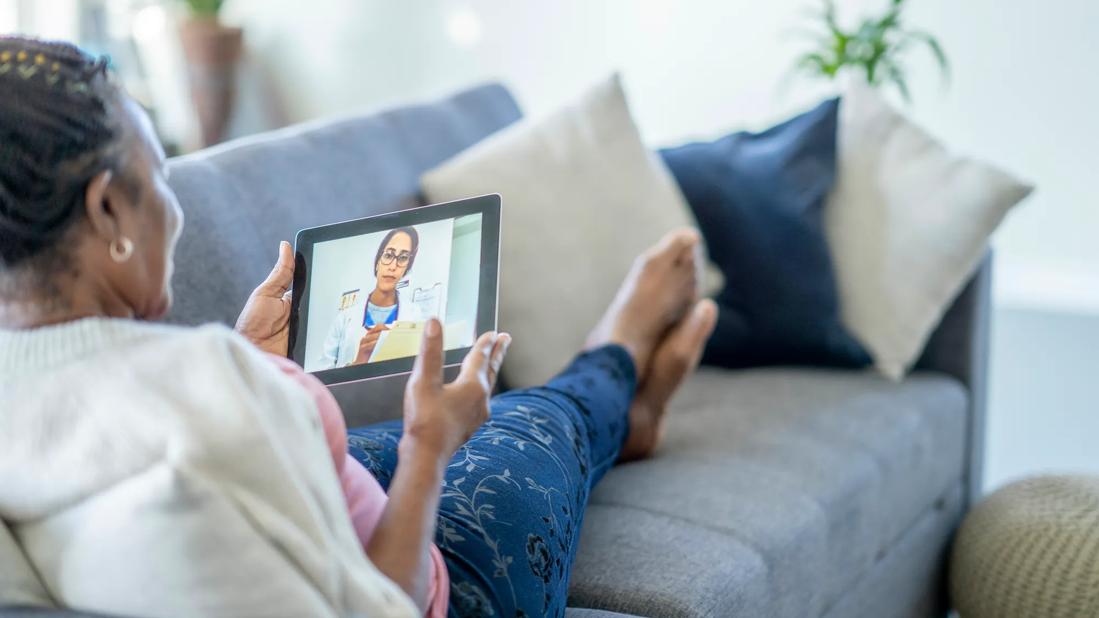 Female reclining on couch holding laptop, having video appointment with healthcare provider