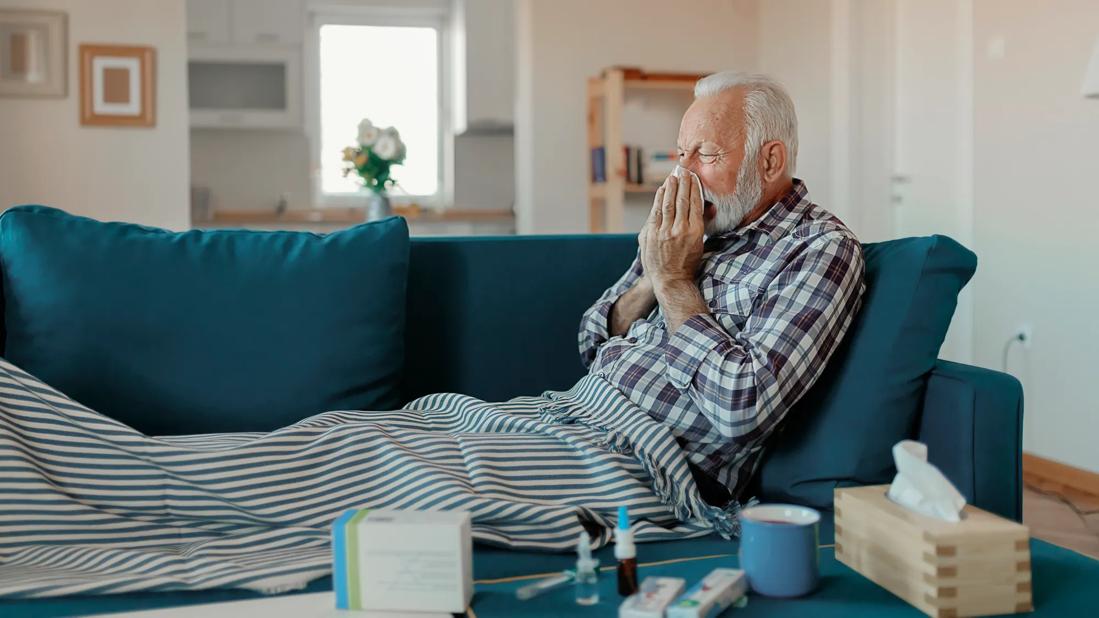 Older male reclining on couch, blowing nose, with cold medications and products on coffee table