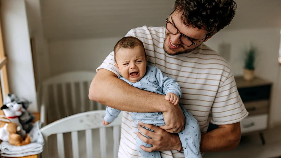 Smiling caregiver holding crying baby in nursery, crib nearby