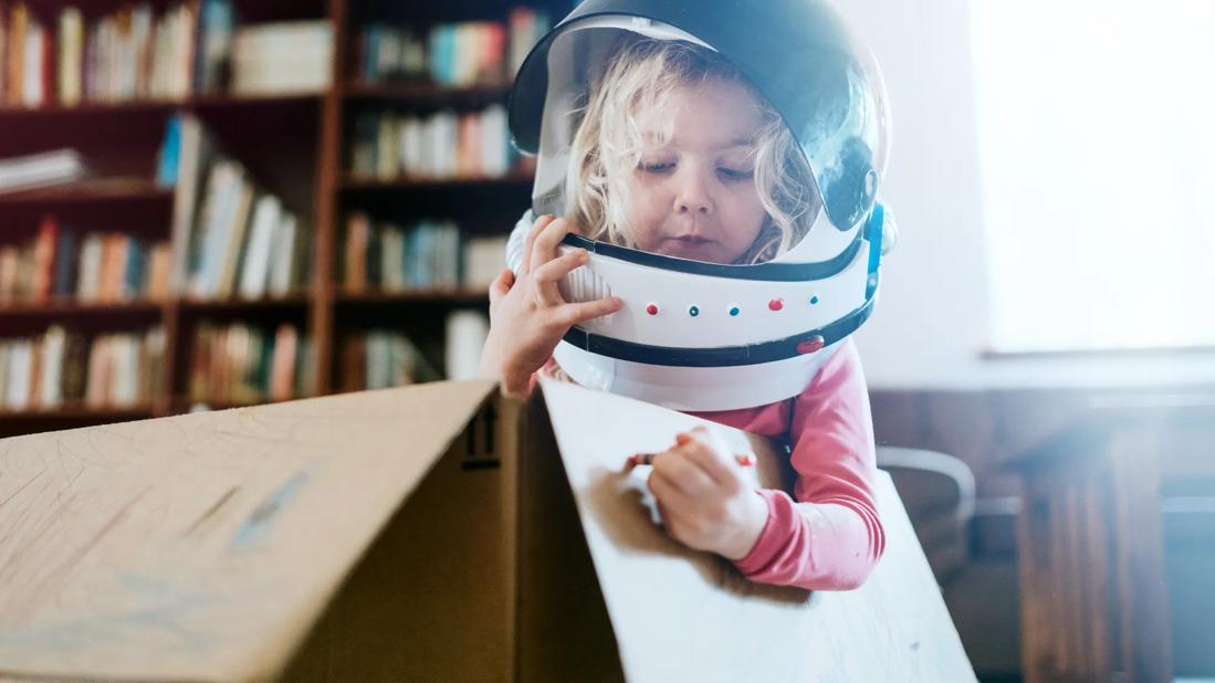 Young child at home, with space helmet on, sitting in cardboard box, coloring on the box