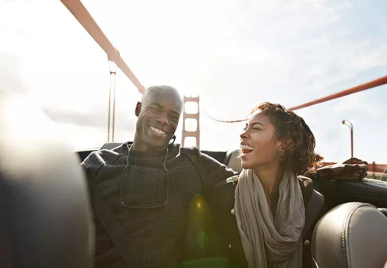 Two smiling adults in a car with a bridge in the background