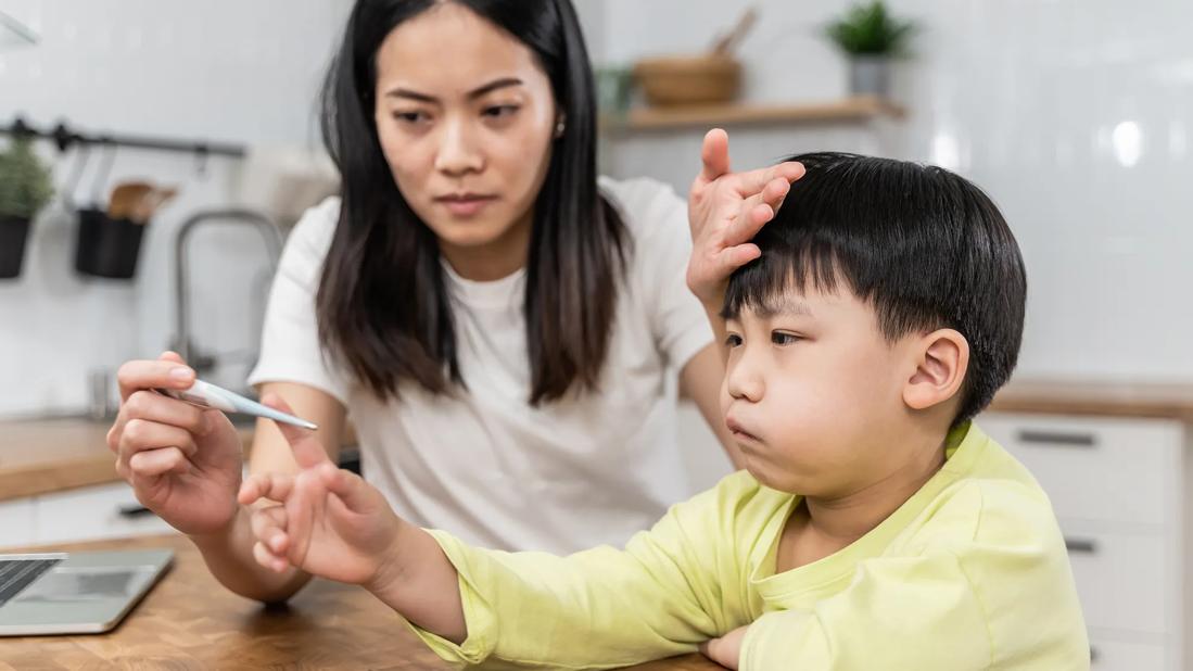 Caretaker reading a digital thermometer, while placing their hand on a sick-looking child's forehead