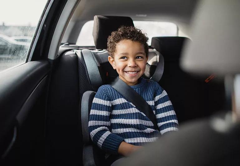 A smiling child strapped into a car seat looks out of a car window.