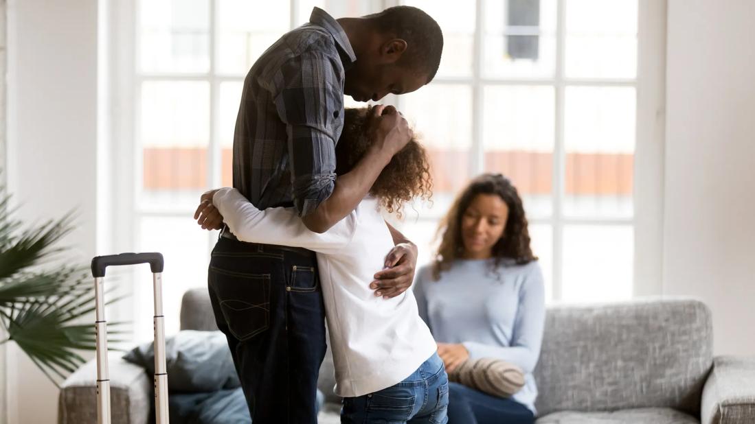 Caregiver standing with suitcase nearby, hugging child, with another caregiver sitting on couch