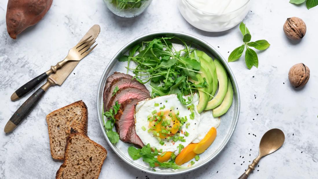 Plate with beef, eggs, avocado, leafy greens and apricots, with multi-grain bread, walnuts, sweet potato and yogurt