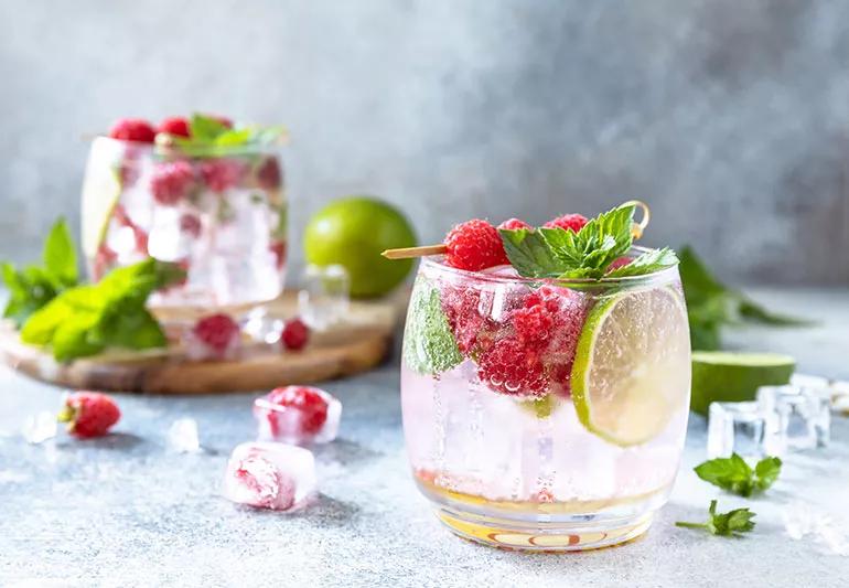 Cold sparkling water with slices of lime, raspberries and mint are in two clear glasses.