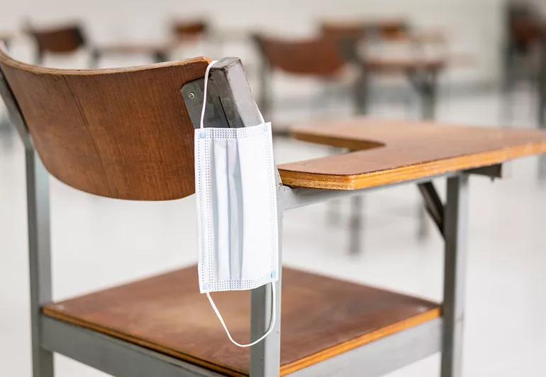 Covid mask hangs on arm of school desk, ready to be used.