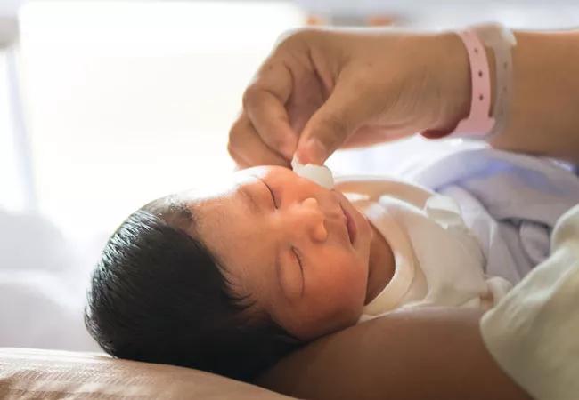 A person gently cradles a newborn baby in their arms while rubbing face with cotton swab.