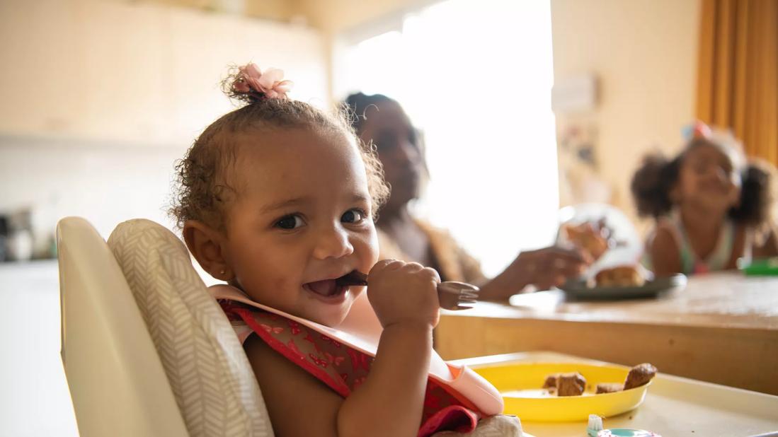 Smiling older baby in high chair with baby utensil in mouth, eating with family