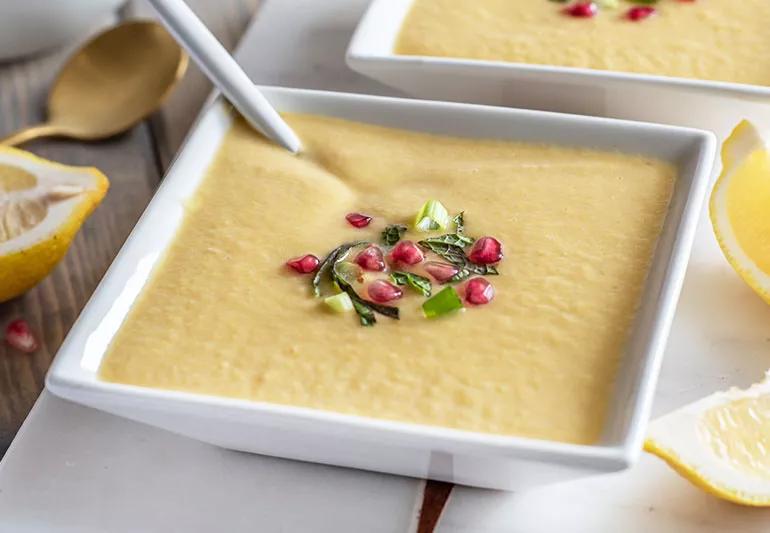 Square bowl of summer squash and chive soup garnished with pomegranate seeds, green onions and herbs