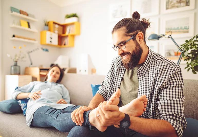 A man with his hair in a bun massaging the feet of a pregnant woman who is laying on a couch