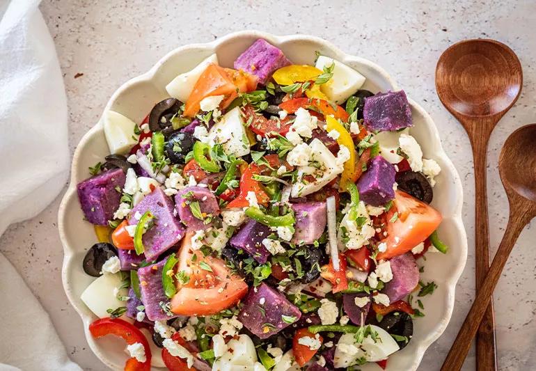 Colorful salad of purple potatoes, tomatoes, feta and greens on a counter next to wooden spoons
