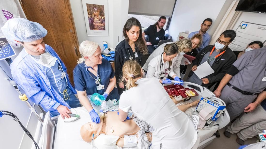 Medical team stands around manikin in hospital bed