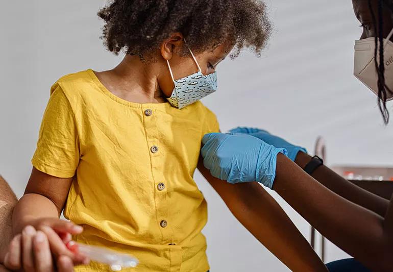 A small, masked child watches as a healthcare provider places a bandaid on their arm after giving them a vaccine.