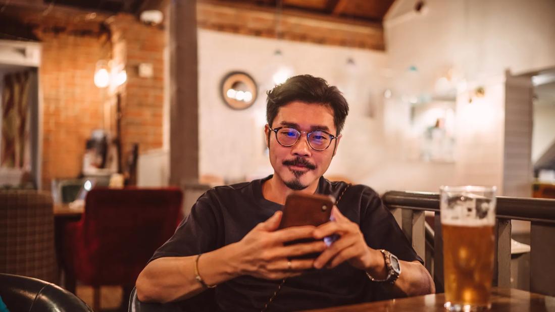 Person sitting at a table in a bar, on their phone, with glass of beer on the table