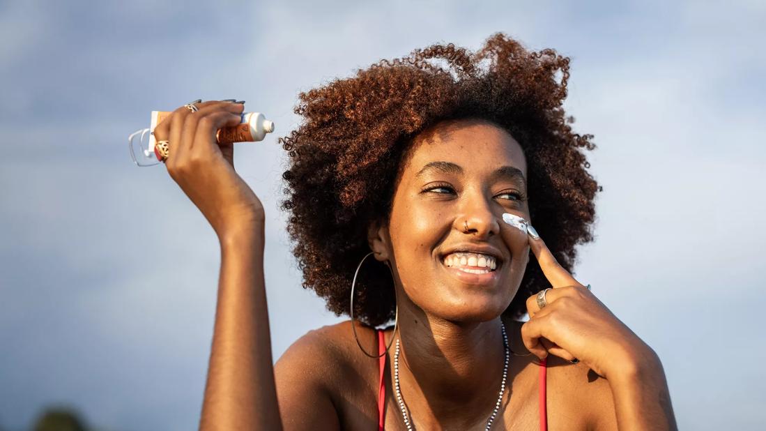 Smiling person under sunny blue sky, holding tube of sunscreen, applying to face