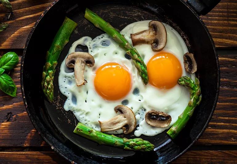 A pair of sunny-side up fried eggs in a skillet with mushrooms and asparagus