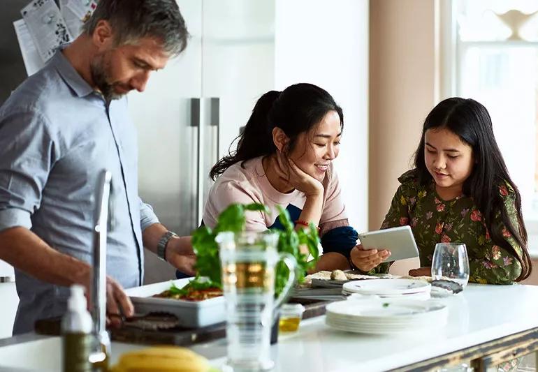 Family gathered at the kitchen counter where Dad is cooking and mom and daughter are talking and checking out online material on an ipad.