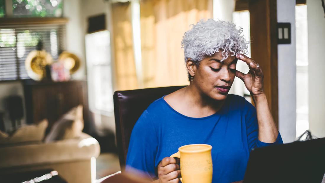 Female looking at laptop at home desk in living room, hand on head, holding coffee cup, looking stressed