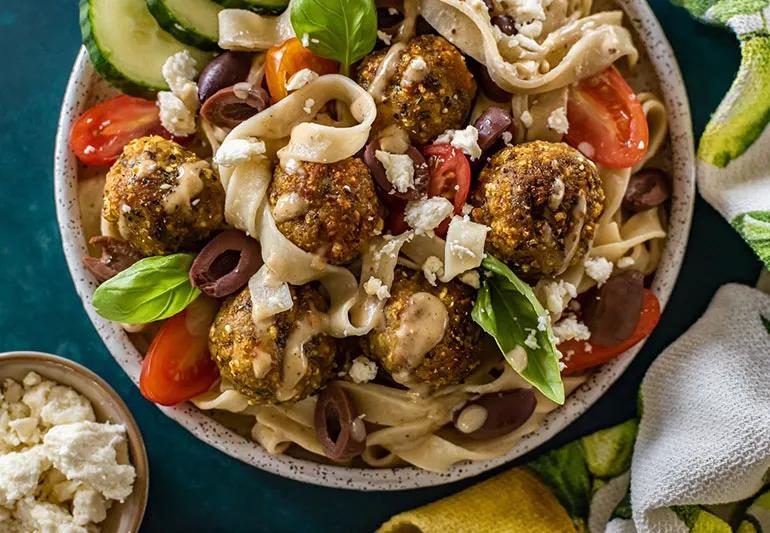 A bowl with noodles, cucumbers, leafy greens, tomatoes and chicken meatballs.