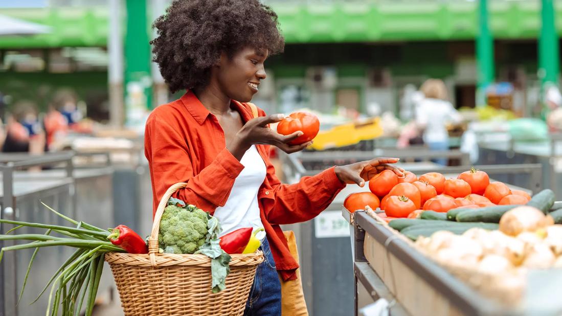 Person in grocery market with basket of fresh veggies picking out tomatoes