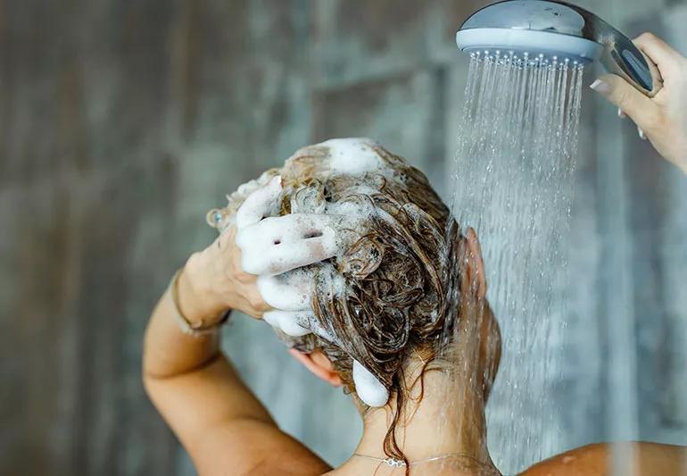 Woman lathering soap into her hair while holding shower head to rinse.