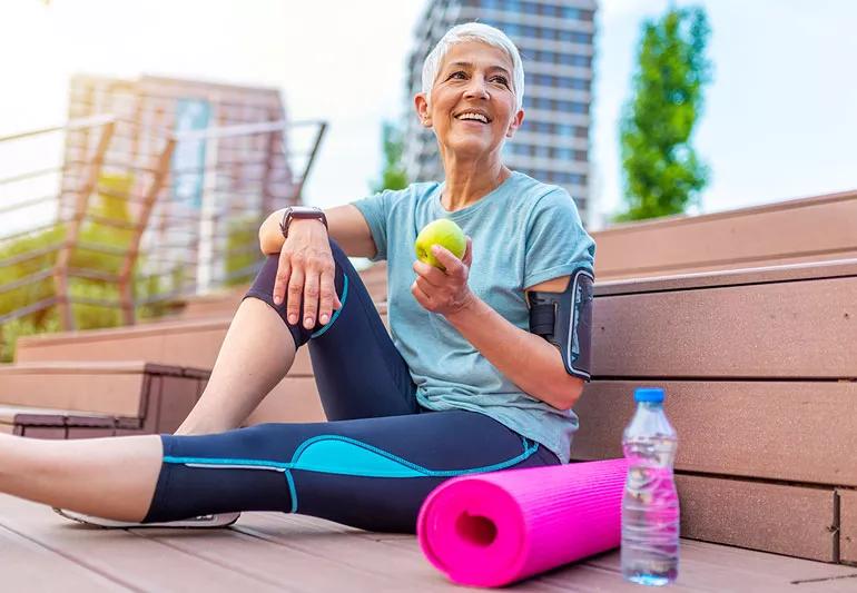 older person in exercise clothes sitting outside next to yoga mat and water bottle holding apple