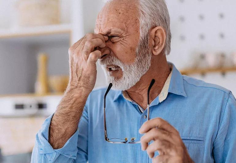 Older person holds eyeglasses while rubbing eyes and squinting.