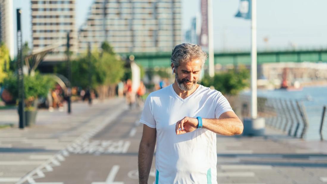 Smiling man walking on paved path outside, looking at smartwatch