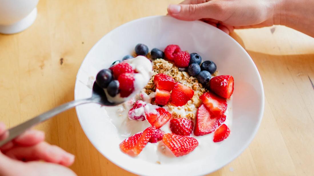 Hand scooping up spoonful of yogurt, granola and fruit from bowl