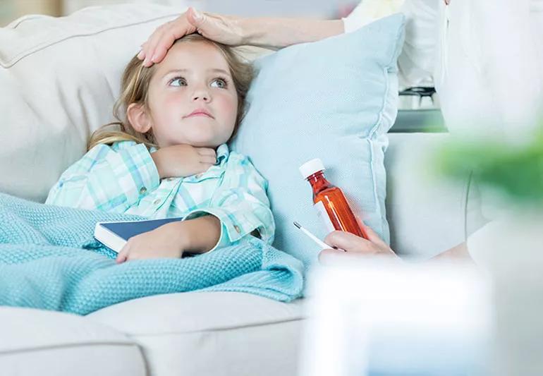 A sick child lying on the couch while an adult feels their forehead for a fever