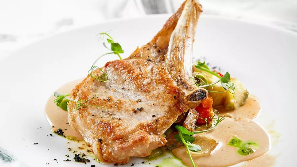 A roasted pork chop on a mound of vegetables with sauce, displayed in a white bowl