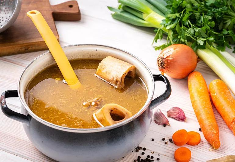 A pot of bone broth with ingredients displayed on a white tabletop in the background.