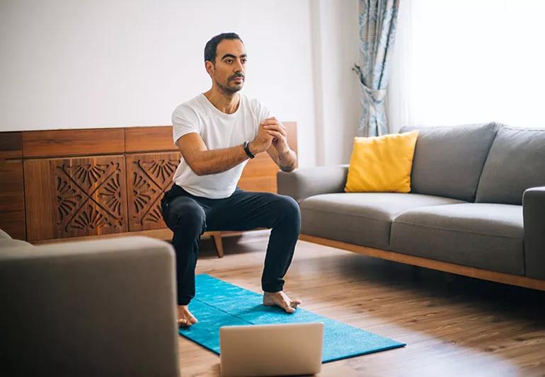 A man in a living room doing squats on a yoga mat in front of a laptop