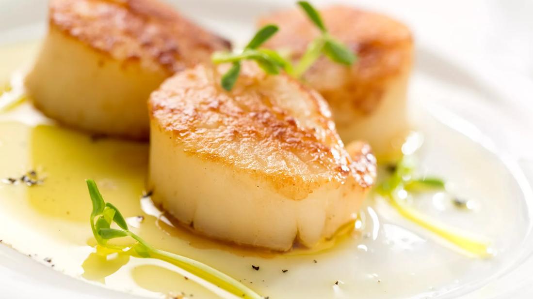 Three large, buttery seared scallops atop olive oil and greens on a white plate