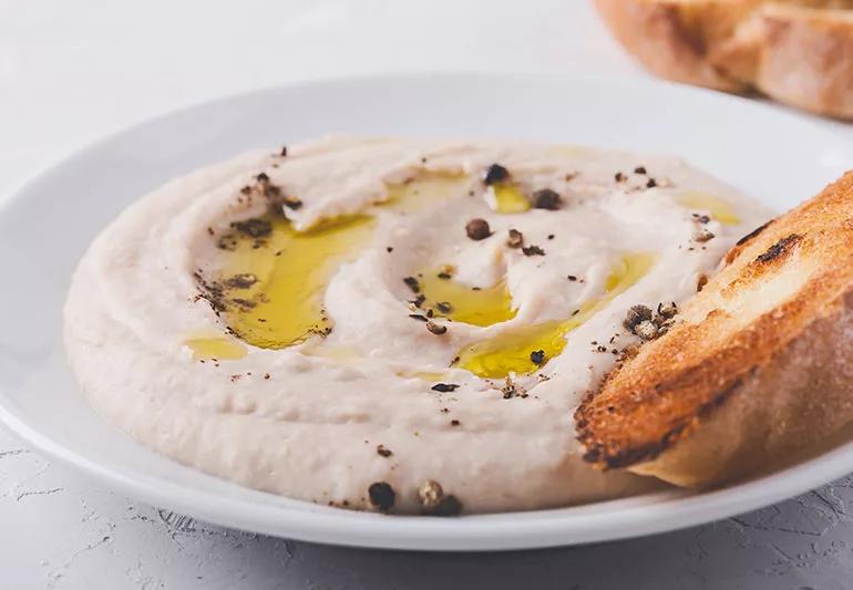 Plate of white bean dip pooled with olive oil and a piece of toasted bread on the side