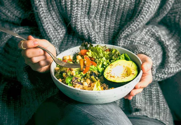 Woman in grey sweater eating a healthy salad filled with greens, beans, tomatoes and an avocado.