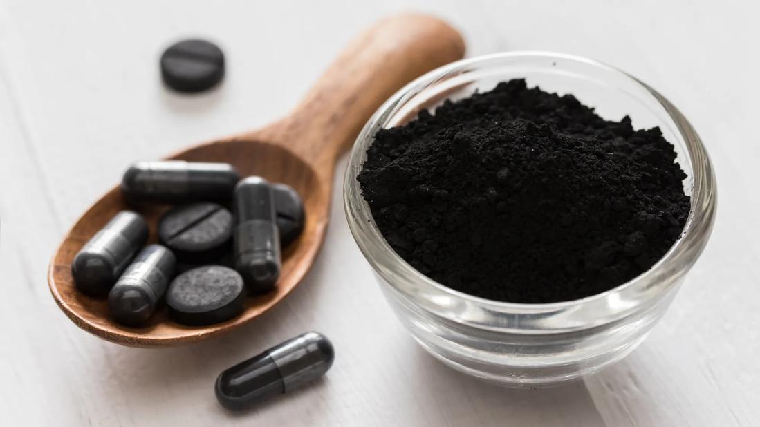 Wooden spoonful of activated charcoal tablets and capsules, and small dish of loose activated charcoal