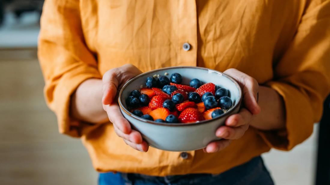 Up close of person's hands holding bowl of strawberries and blueberries over oatmeal